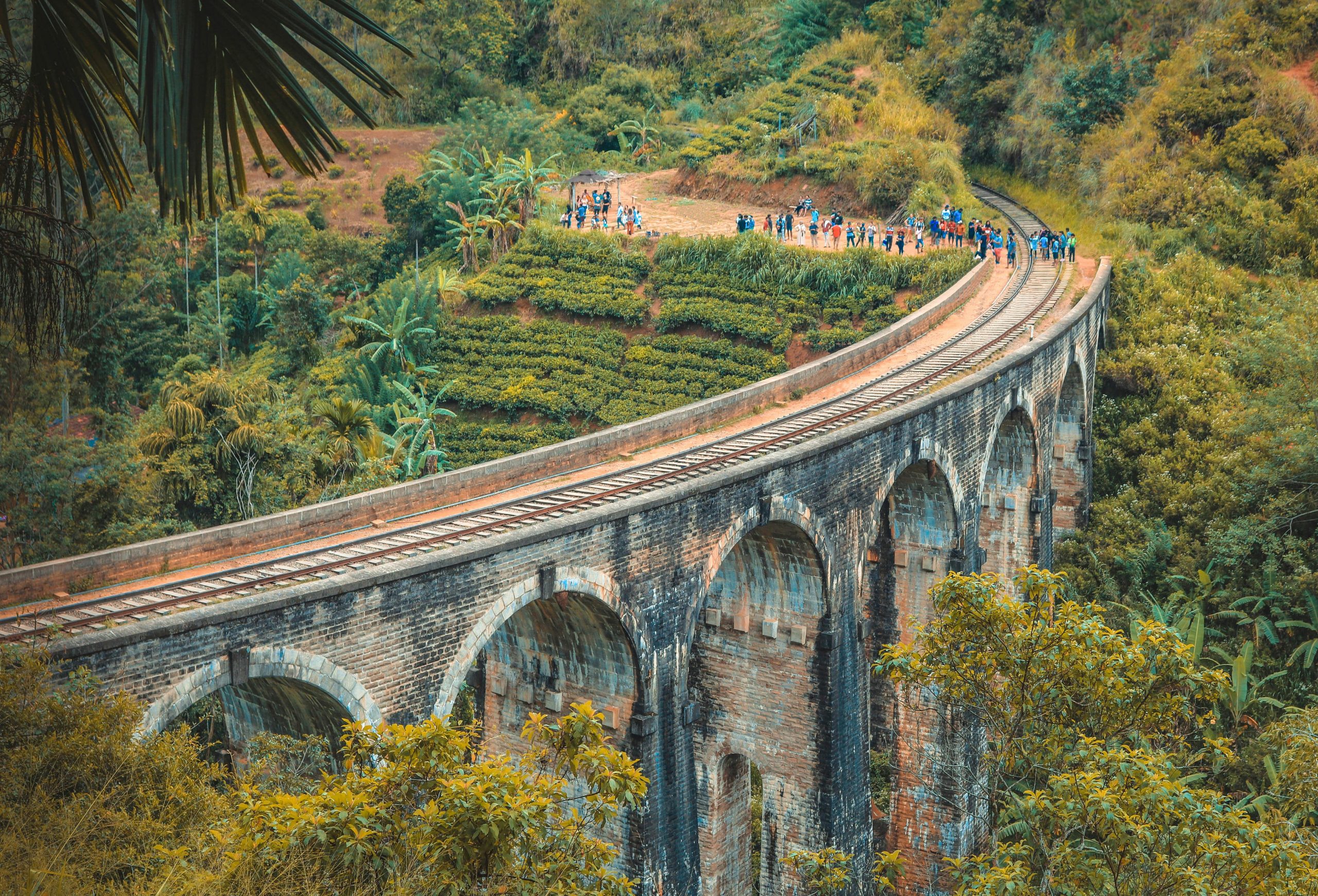 Aerial view of the iconic Nine Arches Bridge surrounded by lush greenery in Ella, Sri Lanka, with tourists enjoying the sight.
