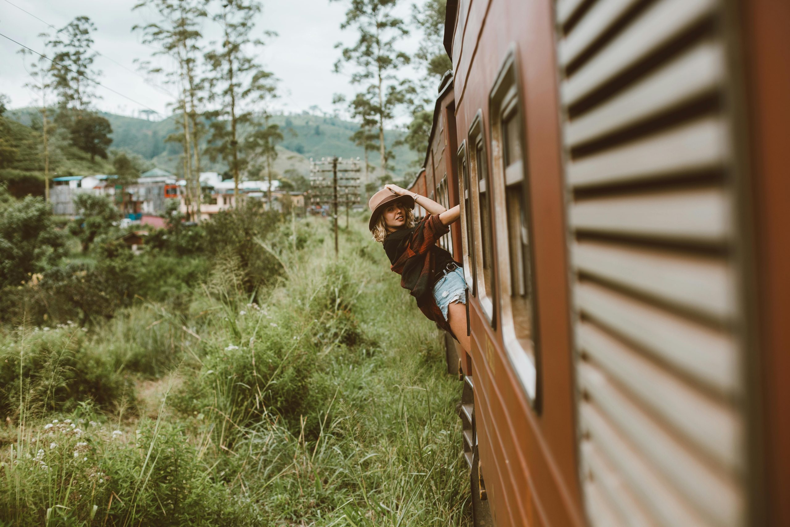 Side view stylish lady in denim shorts standing in cabin door of moving train and touching hat while passing through rural area on summer day